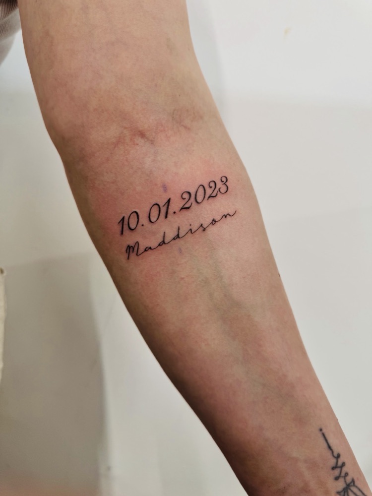 Fine-line script lettering tattoo with a date and name on a forearm